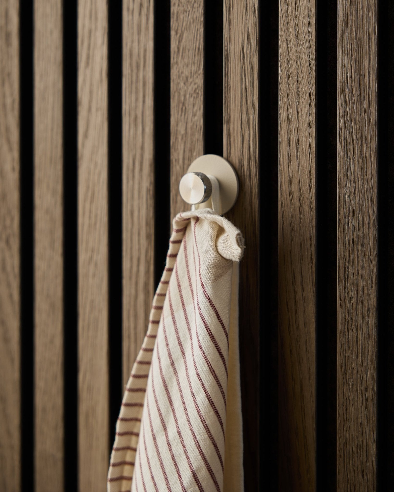 A striped towel hangs on a beige Norr Coat Hook by WoodUpp, made of stainless steel, mounted on a textured wooden Akupanel acoustic slatwall panel.